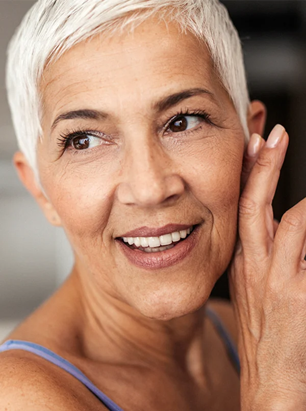 A woman with short white hair, wearing a purple tank top, smiling with one hand pressed against her cheek- Full Mouth Dental Reconsturction in Beverly Hills, CA