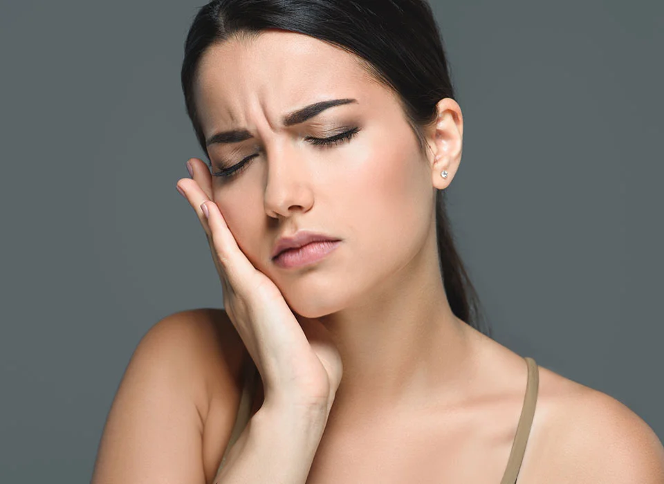 brunette woman, against a grey abckground, touching her jaw in pain - TMJ Treatment in Beverly Hills, CA